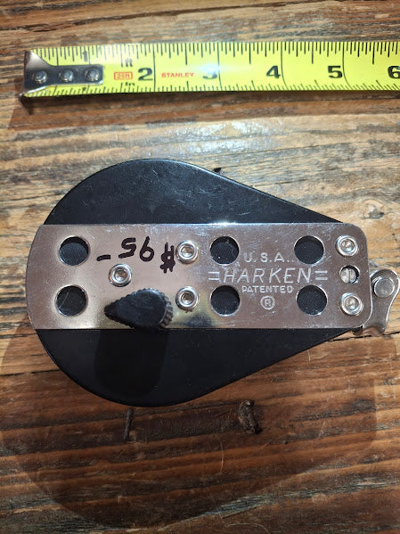 Metal pulley system with 'Harken' branding on a wooden surface, measured by a tape measure.