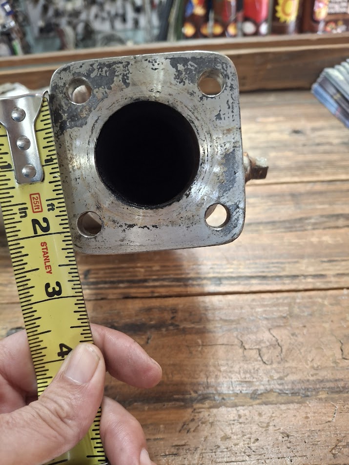 Metal exhaust pipe, held by a hand with a tape measure for scale, on a wooden surface.